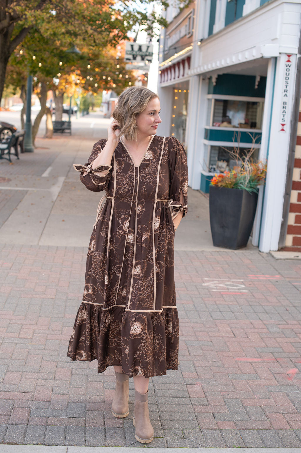 Woman in a brown patterned dress standing on a sidewalk with shops in the background
