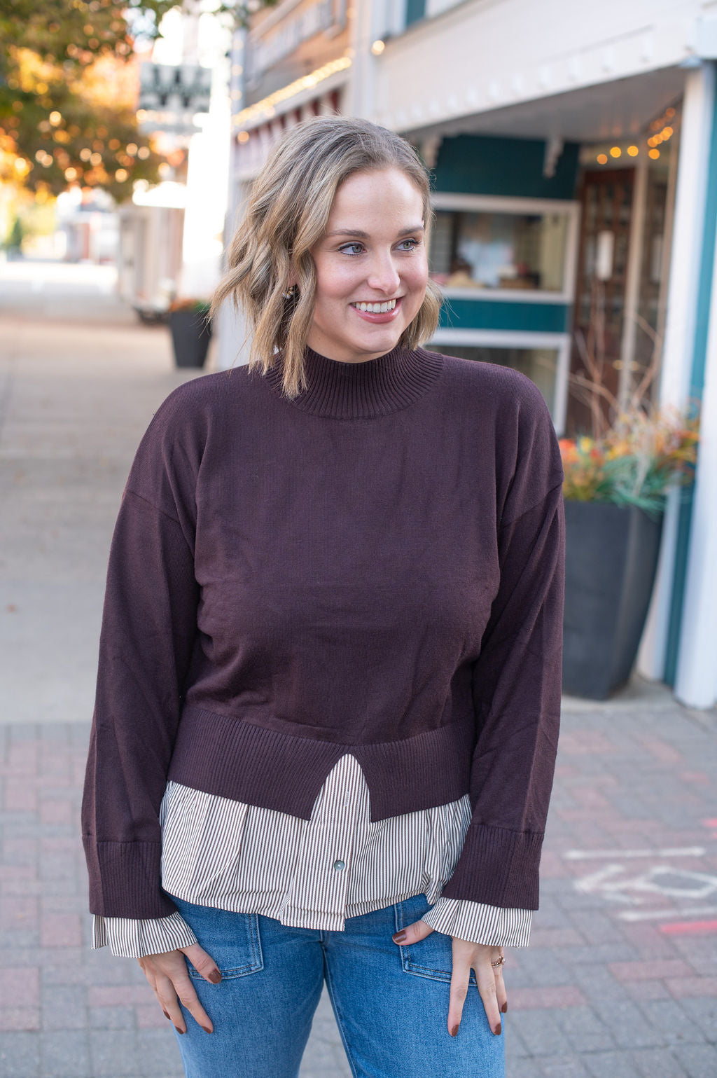Woman wearing chocolate brown sweater over a striped shirt with blue jeans, standing on a sidewalk.