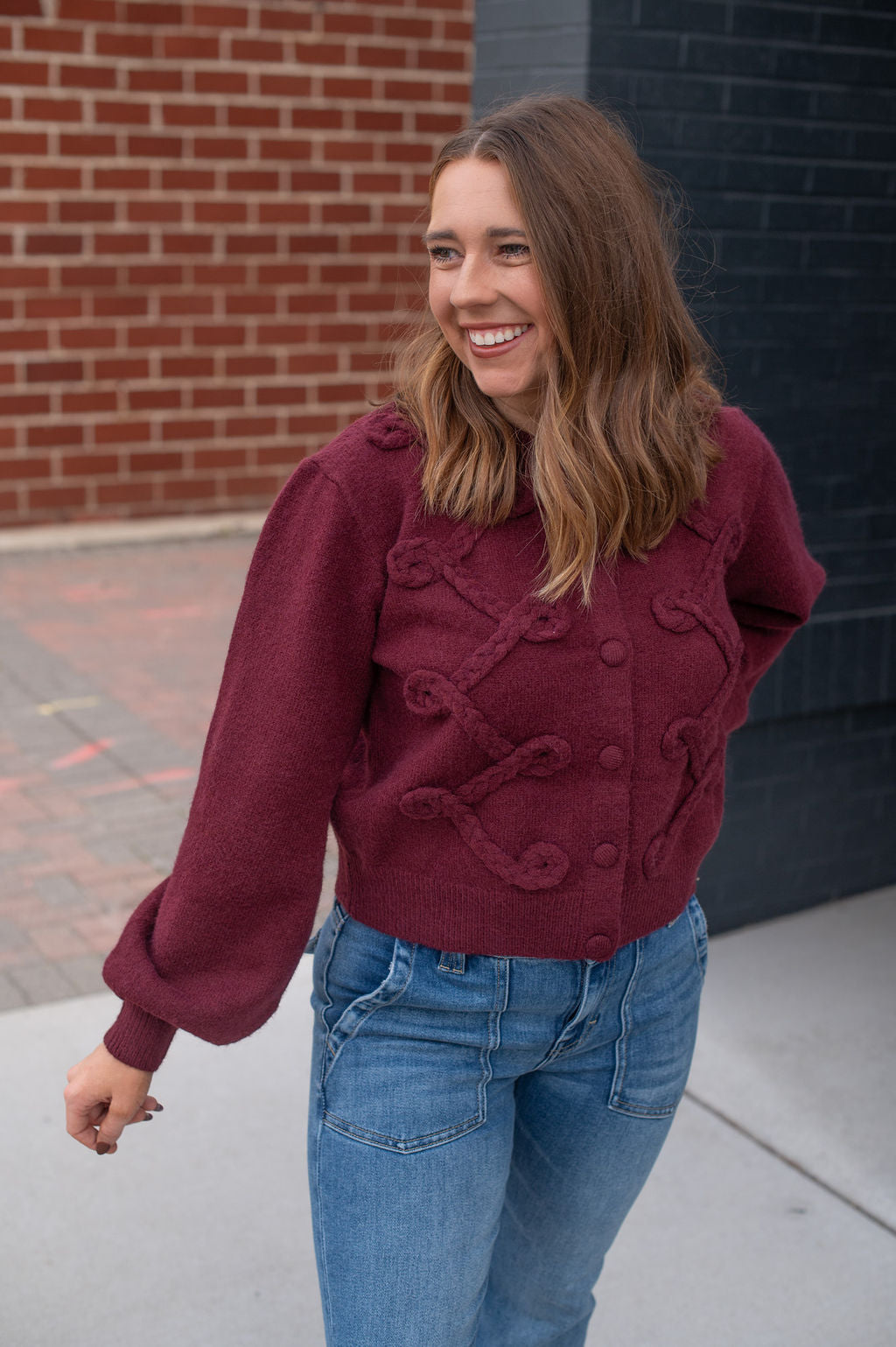Woman wearing a maroon sweater with button details and blue jeans, standing against a brick wall.