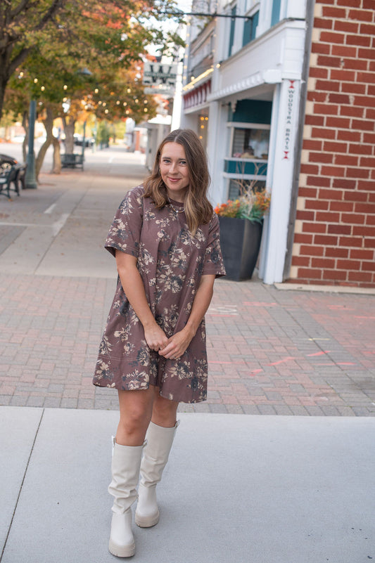 Woman in a floral dress and white boots standing on a sidewalk with a building and trees in the background.
