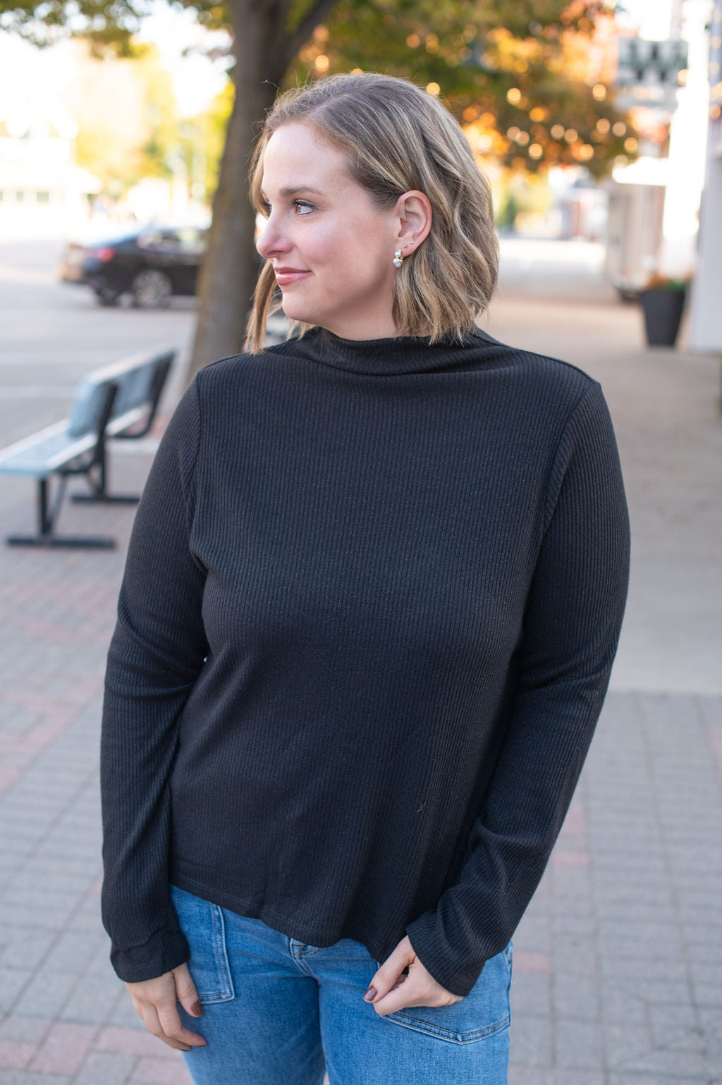 Woman wearing a black sweater and blue jeans standing on a sidewalk.