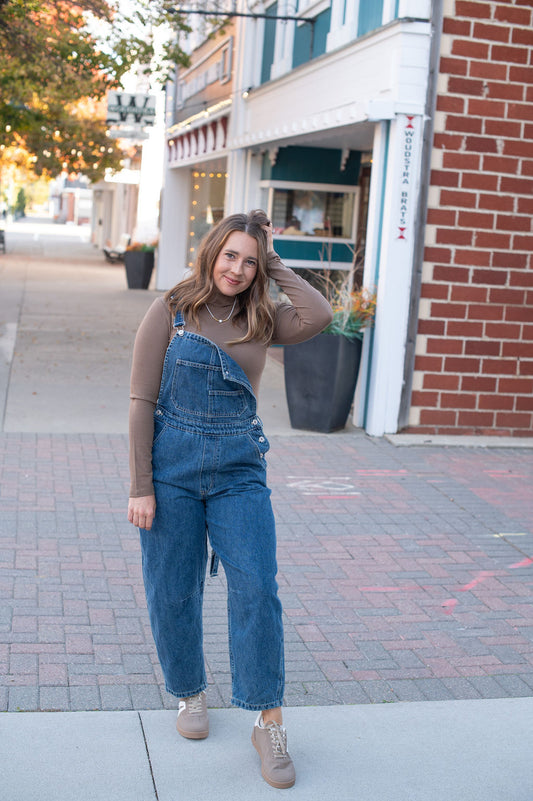 Woman wearing blue denim overalls standing on a sidewalk with buildings in the background