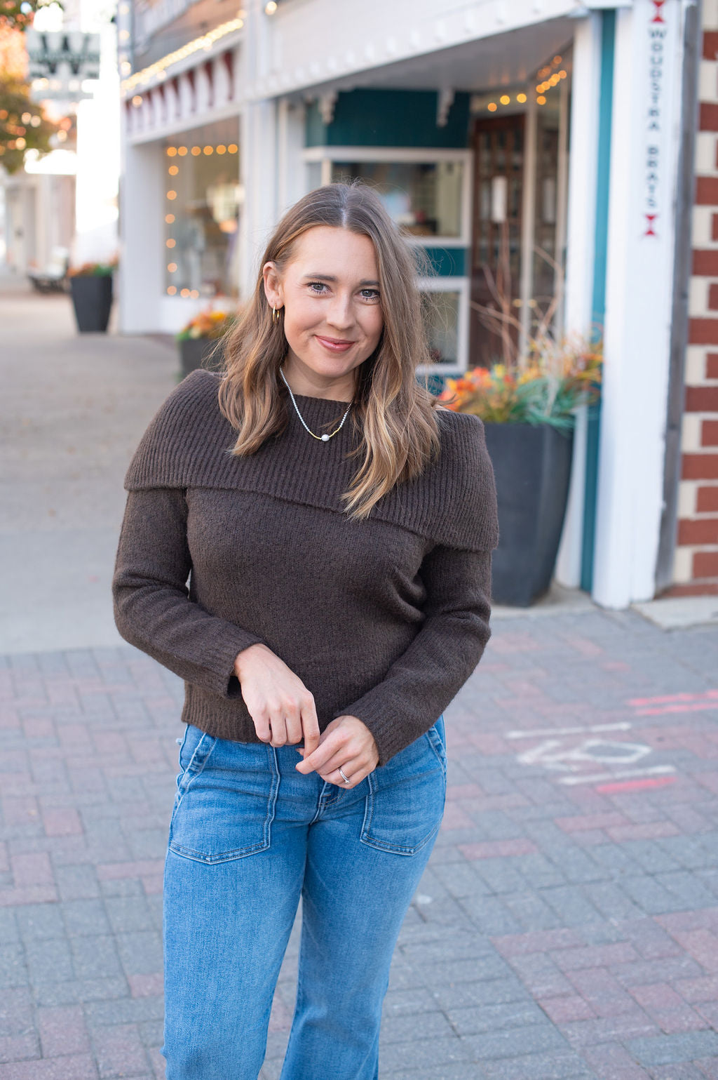 Woman wearing a brown sweater and blue jeans standing on a sidewalk with storefronts in the background.