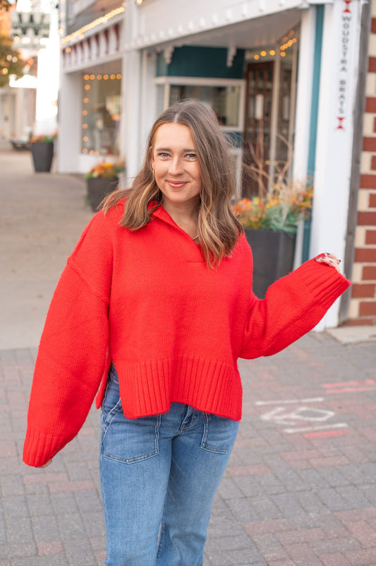 Woman wearing a red sweater and blue jeans standing on a sidewalk with storefronts in the background.