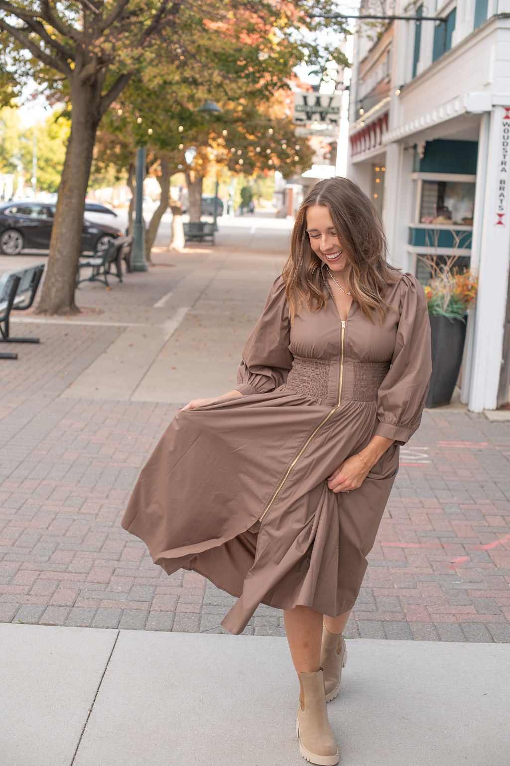 Woman in a brown dress standing on a sidewalk with trees and buildings in the background