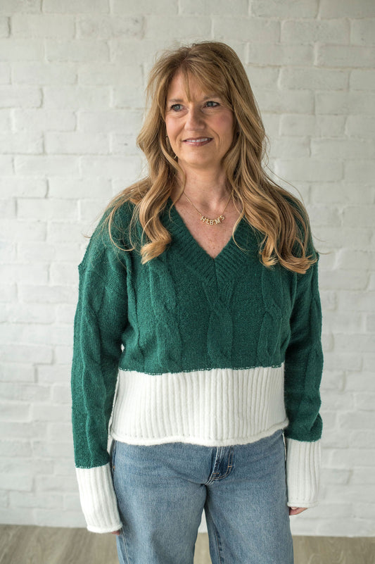 Woman wearing a green sweater with white trim and blue jeans against a white brick wall.