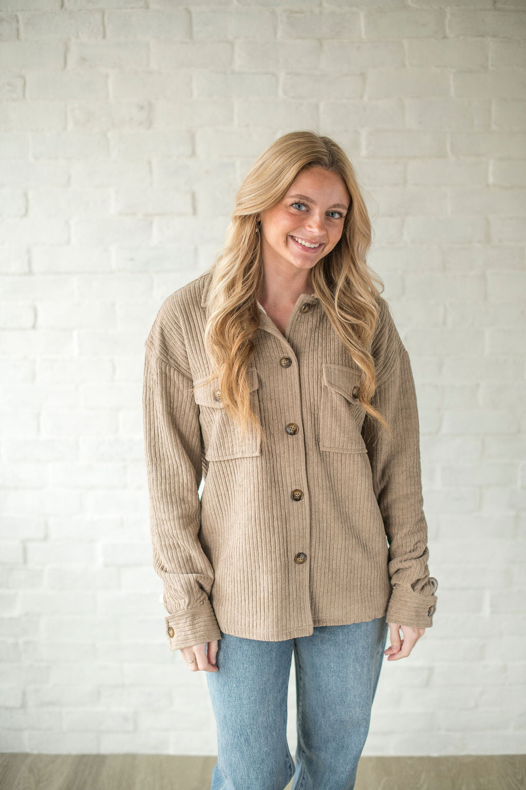 Woman wearing a beige corduroy jacket and blue jeans against a white brick wall.