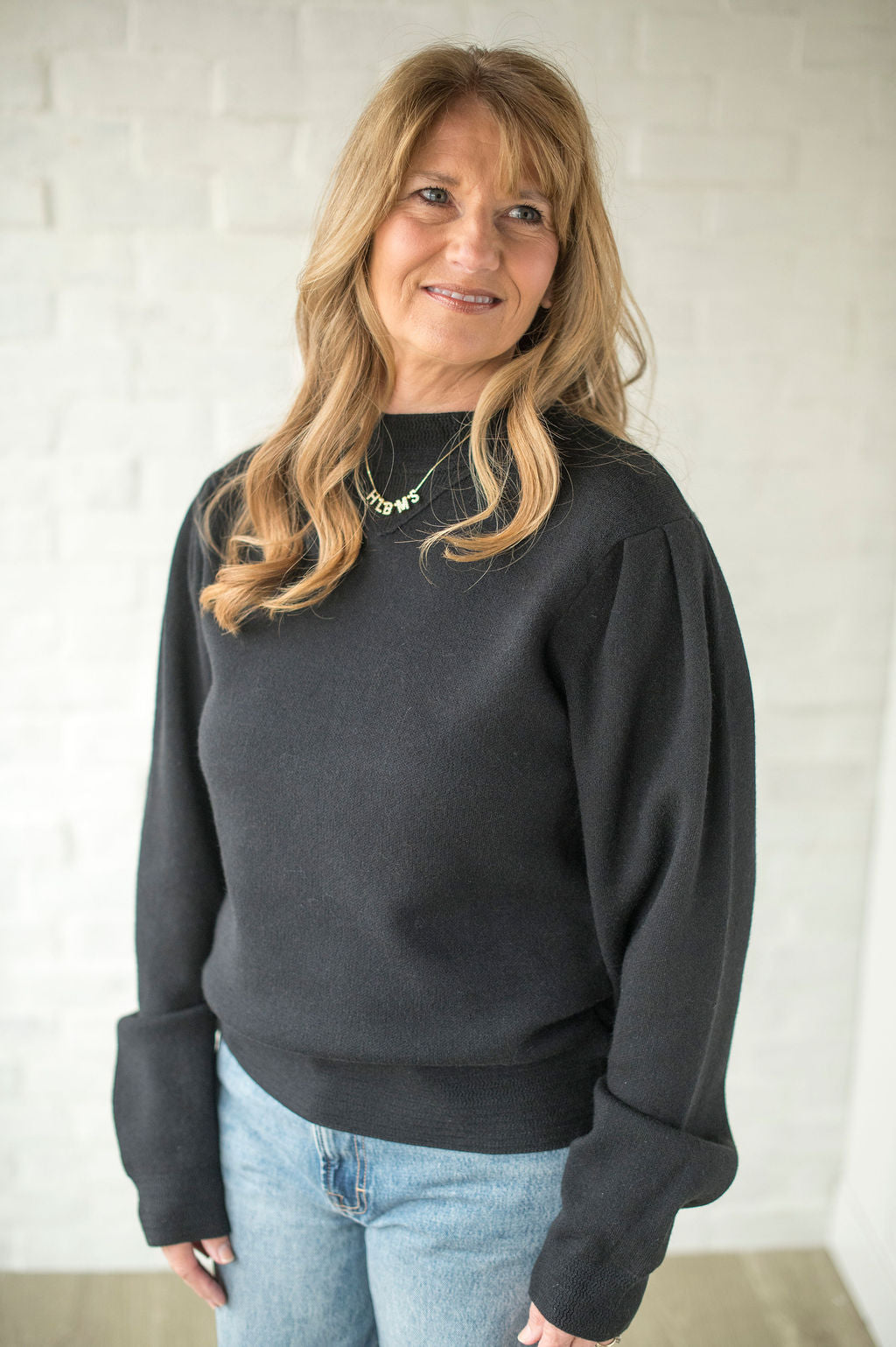 Woman wearing a dark gray sweatshirt and light blue jeans against a white brick wall.