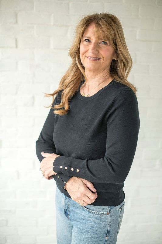 Woman wearing a dark black sweater with decorative buttons, standing against a white brick wall.