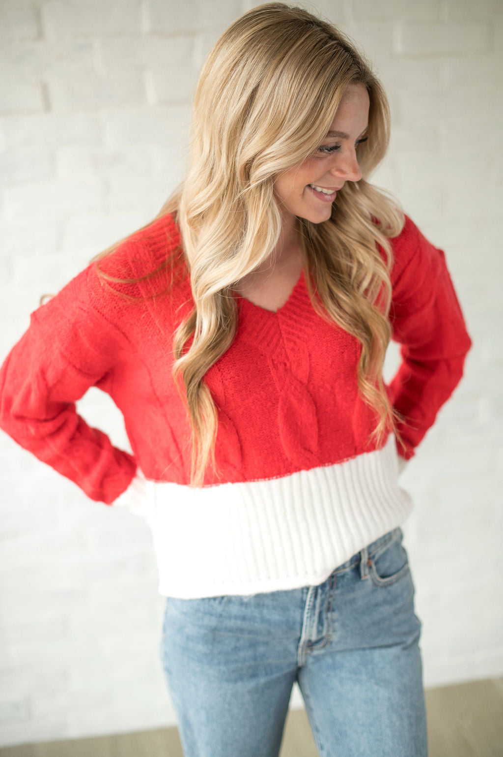 Woman wearing a red and white sweater with blue jeans against a white brick wall.