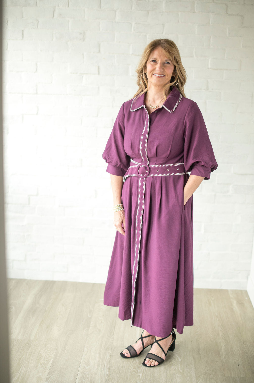 Woman wearing a purple dress with white trim against a white brick wall.