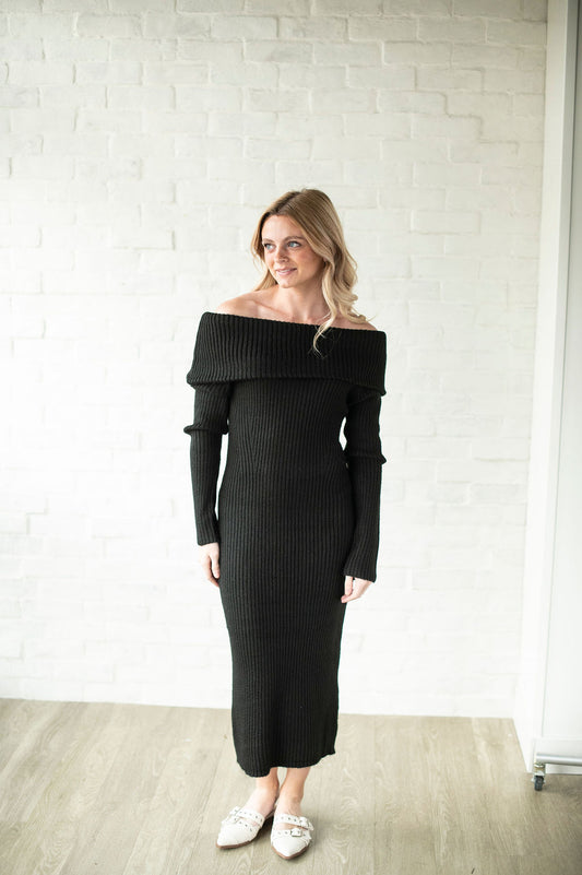Woman wearing a black off-shoulder dress against a white brick wall.